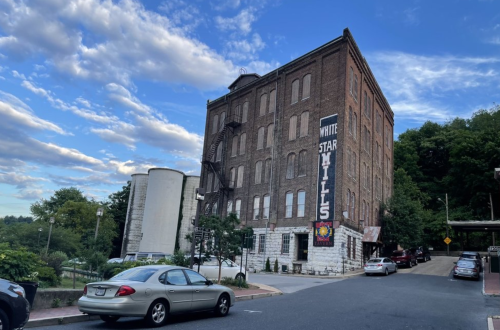 Historic brick building with "White Star Mills" sign, surrounded by trees and parked cars under a cloudy blue sky.