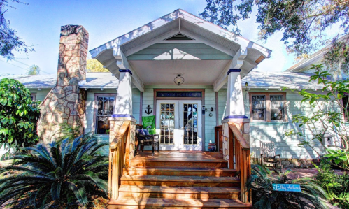 A charming house with a wooden porch, stone chimney, and lush greenery surrounding the entrance.