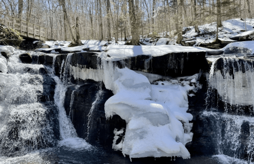 A frozen waterfall surrounded by snow-covered rocks and trees in a winter landscape.