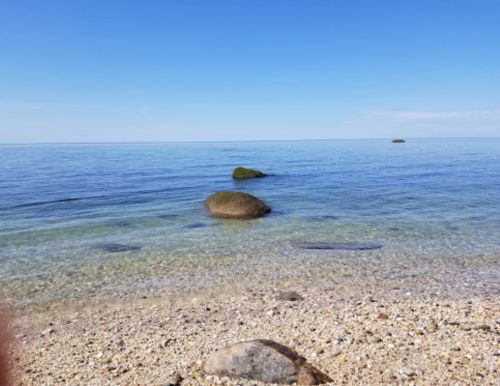 Calm blue waters with smooth rocks visible, sandy beach in the foreground, under a clear blue sky.