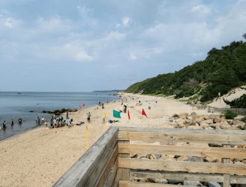 A sandy beach with people enjoying the water, colorful flags, and rocky areas under a partly cloudy sky.