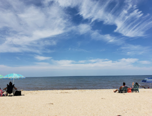 A sandy beach with a calm sea, two people sitting on chairs, and colorful umbrellas under a blue sky with clouds.