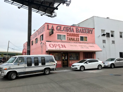 A pink bakery building with "La Gloria Bakery" sign, featuring an open banner and parked cars outside.