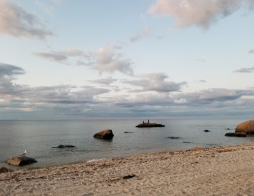 A serene beach scene with gentle waves, rocks, and a cloudy sky, featuring two people on a distant rock.