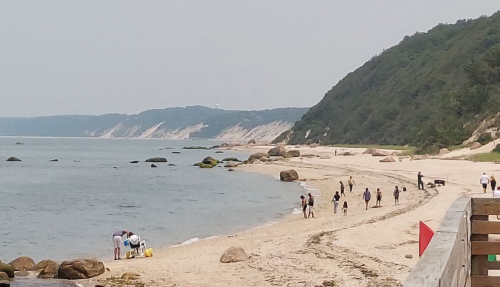 A sandy beach with people walking, gathering near the water, and rocky formations along a scenic coastline.