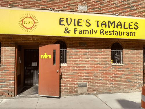 A brick building with a yellow sign reading "Evie's Tamales & Family Restaurant" and a brown door slightly open.