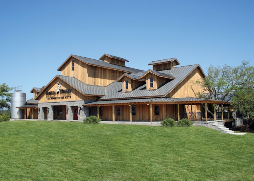 A large wooden winery building with a metal roof, surrounded by green grass and clear blue skies.