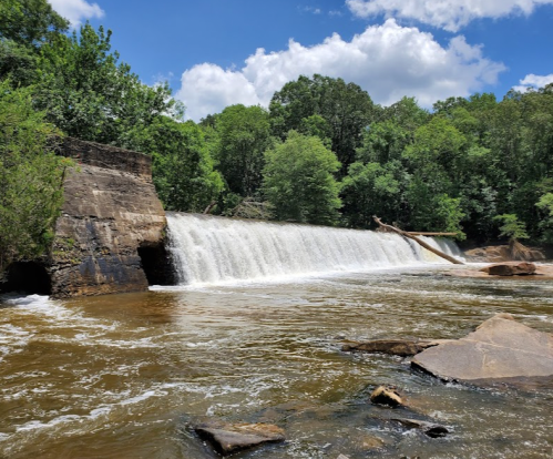 A serene waterfall cascades over a stone dam, surrounded by lush green trees and a clear blue sky.