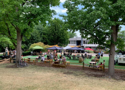 A lively outdoor gathering with people sitting at tables under tents, surrounded by trees and a grassy area.