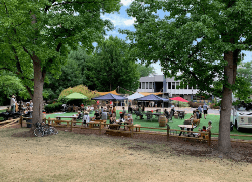 A lively outdoor gathering with people sitting at tables under tents, surrounded by trees and a grassy area.