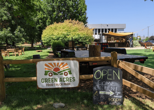 A sunny food truck park with a sign reading "Green Acres," featuring picnic tables and a welcoming "Open" chalkboard sign.