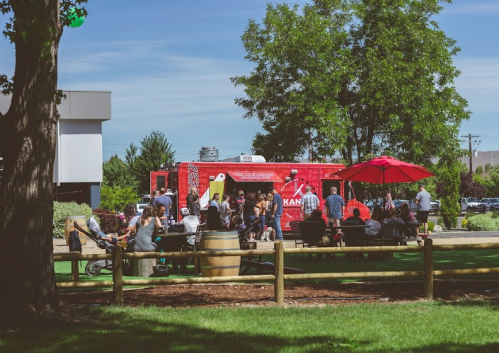 A vibrant food truck in a park, with people enjoying food and drinks under umbrellas and trees.