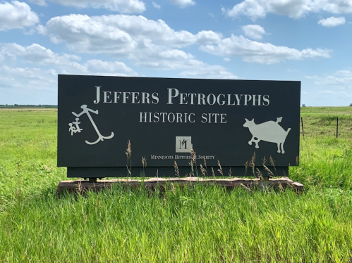 Sign for Jeffers Petroglyphs Historic Site, featuring petroglyph symbols, surrounded by green grass and a blue sky.