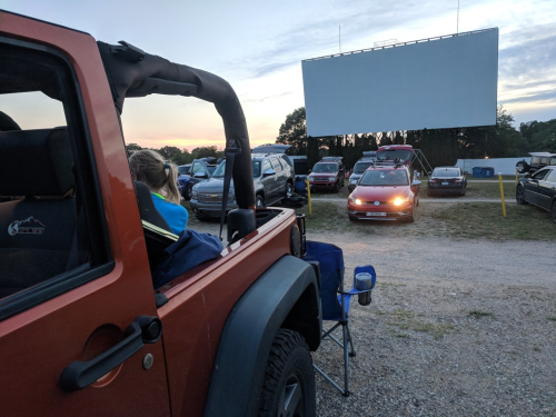 A drive-in movie scene with cars parked, a large screen in the background, and a person sitting in a Jeep.