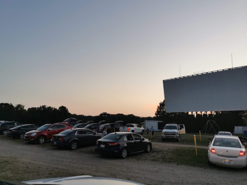 Cars parked at a drive-in theater with a large blank screen and a sunset in the background.