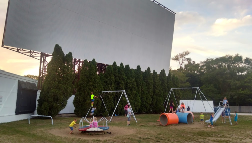 Children play on a playground with swings, a slide, and a merry-go-round, set against a large outdoor screen.