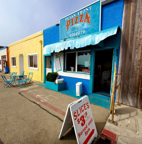 A colorful pizza shop with a blue and yellow exterior, featuring an open sign and a sandwich board advertising pizza slices.