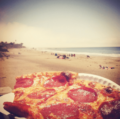 A slice of pepperoni pizza in the foreground with a sunny beach and people in the background.