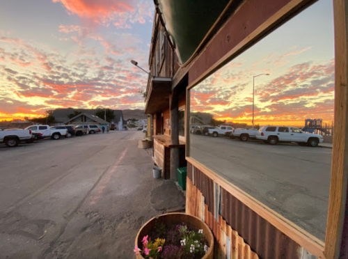 A sunset reflects in a window, with parked cars and a rustic building in the foreground.