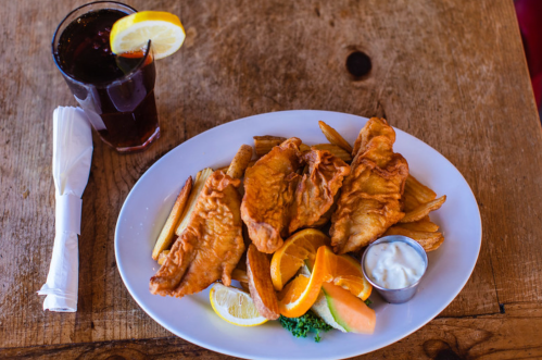 A plate of fried fish with fries, lemon slices, and a small cup of sauce, alongside a glass of iced tea with lemon.