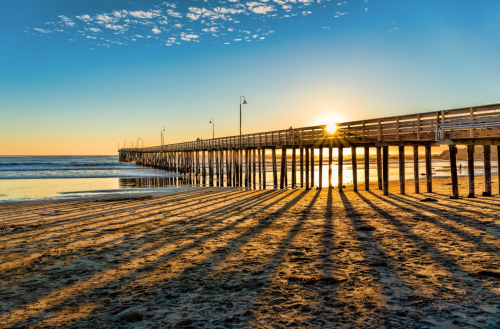 A wooden pier extends into the ocean at sunset, casting long shadows on the sandy beach.