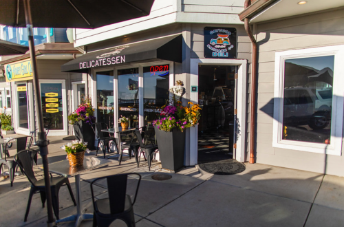 A deli storefront with an "Open" sign, surrounded by colorful flowers and outdoor seating.