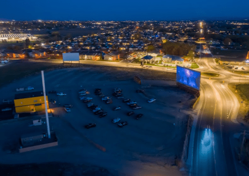 Aerial view of a drive-in theater at night, with parked cars and illuminated buildings in the background.