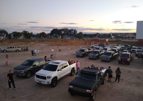 A gathering of trucks in a dirt lot at sunset, with people socializing and a playground in the background.