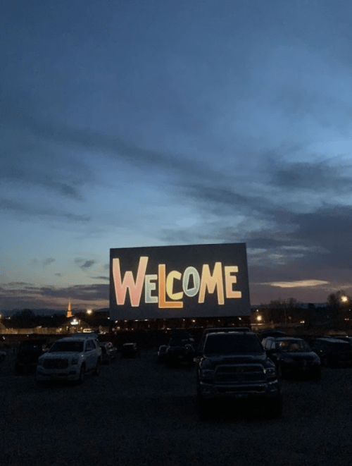 A large outdoor screen displays the word "WELCOME" against a twilight sky, with parked cars in the foreground.