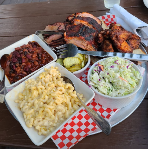 A platter of barbecue featuring smoked meats, macaroni and cheese, baked beans, coleslaw, and pickles on a checkered tray.