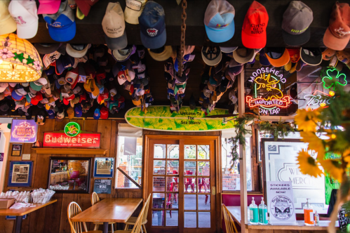 A cozy bar interior with a variety of hats hanging from the ceiling, neon signs, and wooden decor.