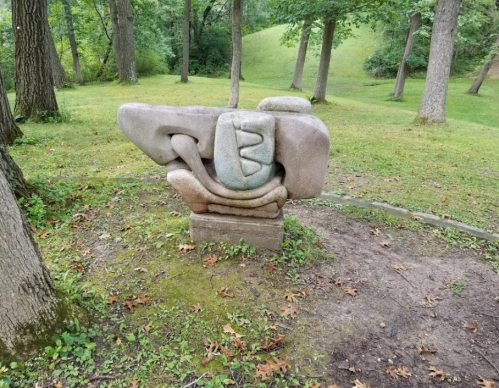 A large abstract stone sculpture in a grassy park, surrounded by trees and a gentle hillside.
