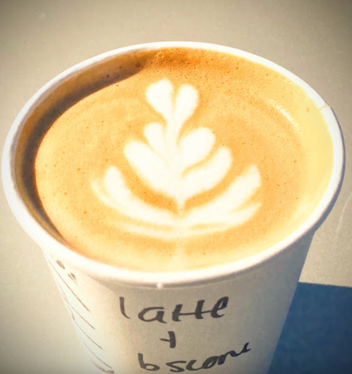 A close-up of a latte in a paper cup, featuring a decorative leaf pattern in the foam.