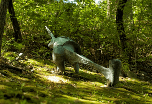 A dinosaur sculpture in a forest, partially covered in moss, surrounded by green foliage and trees.