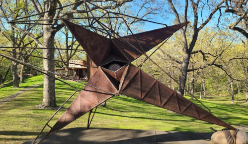 A large, abstract metal sculpture with a star-like shape, set in a green park with trees and a building in the background.