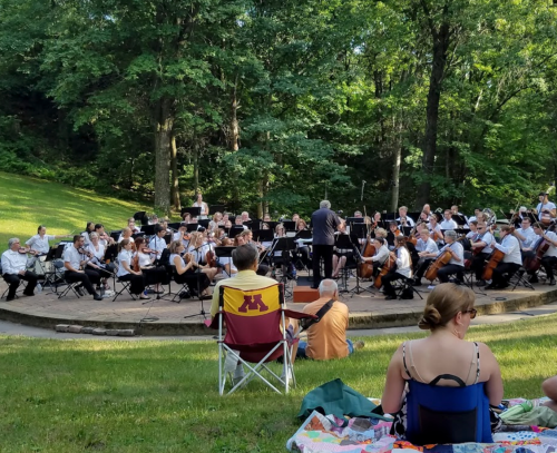 An outdoor orchestra performs in a park, with audience members seated on the grass, surrounded by trees.