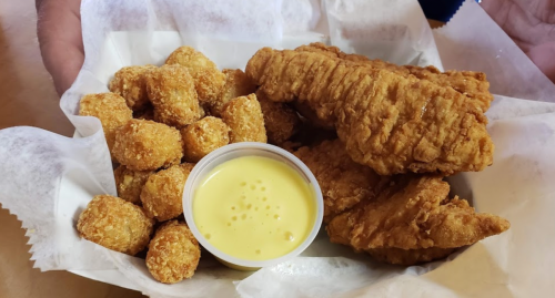 A basket of crispy fried chicken strips and golden tater tots, served with a small cup of dipping sauce.