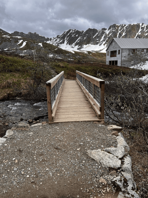 A wooden bridge spans a stream, leading to a rustic cabin surrounded by mountains and snow-capped peaks.