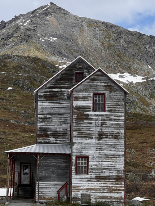 A weathered wooden house with a steep roof, set against a mountainous landscape with patches of snow.