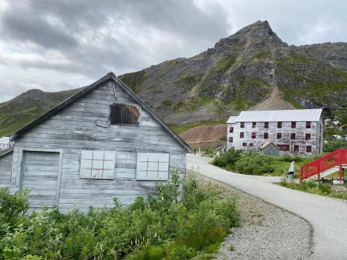 A weathered wooden building and a larger structure on a path, with a mountain backdrop under a cloudy sky.