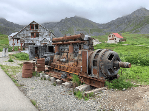 An old, rusted machine sits beside a gravel path, with mountains and historic buildings in the background.