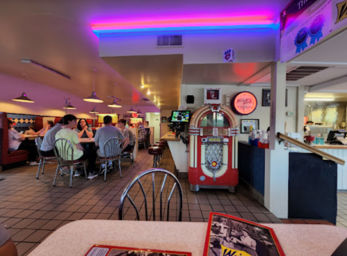 A retro diner interior with colorful lighting, a jukebox, and patrons seated at tables.