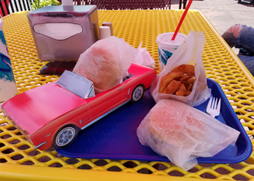 A red toy car on a tray with wrapped burgers, onion rings, and a drink, set on a yellow outdoor table.