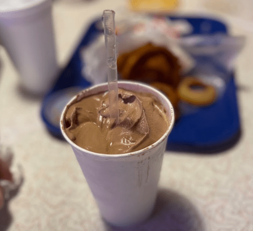 A cup of chocolate soft serve with a clear straw, placed on a tray with food in the background.