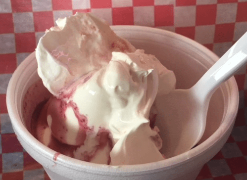 A cup of ice cream with swirls of red sauce and a white spoon, set against a checkered red and white background.