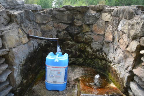 A blue water container sits next to a stone wall with a water spout, surrounded by natural greenery.
