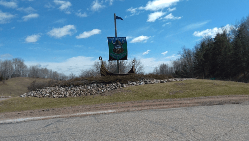 A large Viking ship sculpture with a flag, surrounded by grass and rocks, under a blue sky with scattered clouds.