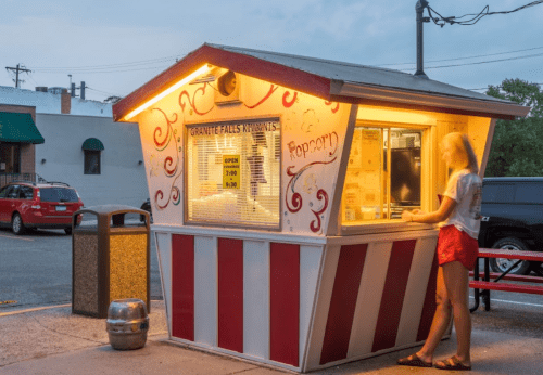 A brightly lit popcorn stand with red and white stripes, featuring a person ordering at dusk.