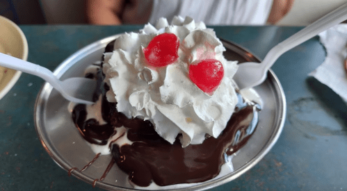 A dessert with chocolate sauce, whipped cream, and two cherries on top, served in a silver dish with two spoons.