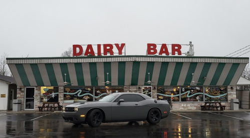 A gray car parked in front of a retro-style dairy bar with green and white striped awning on a cloudy day.